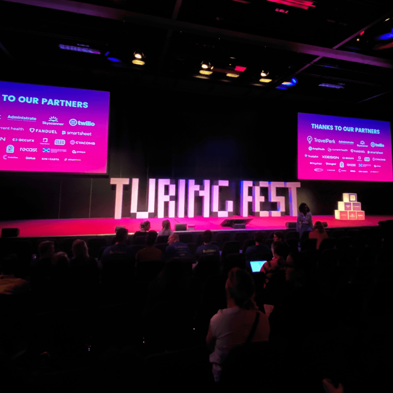 Turing Fest stage one. Two large screens showing all the sponsors. Large white blocks in the centre of the stage spelling Turing Fest